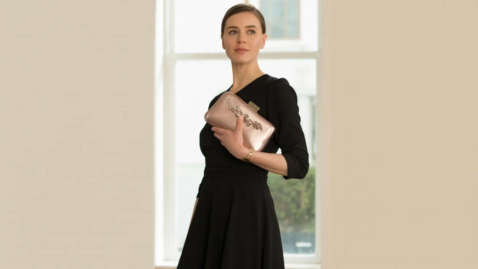 A woman wearing a black dress holds a clutch bag, posing elegantly against a neutral background.