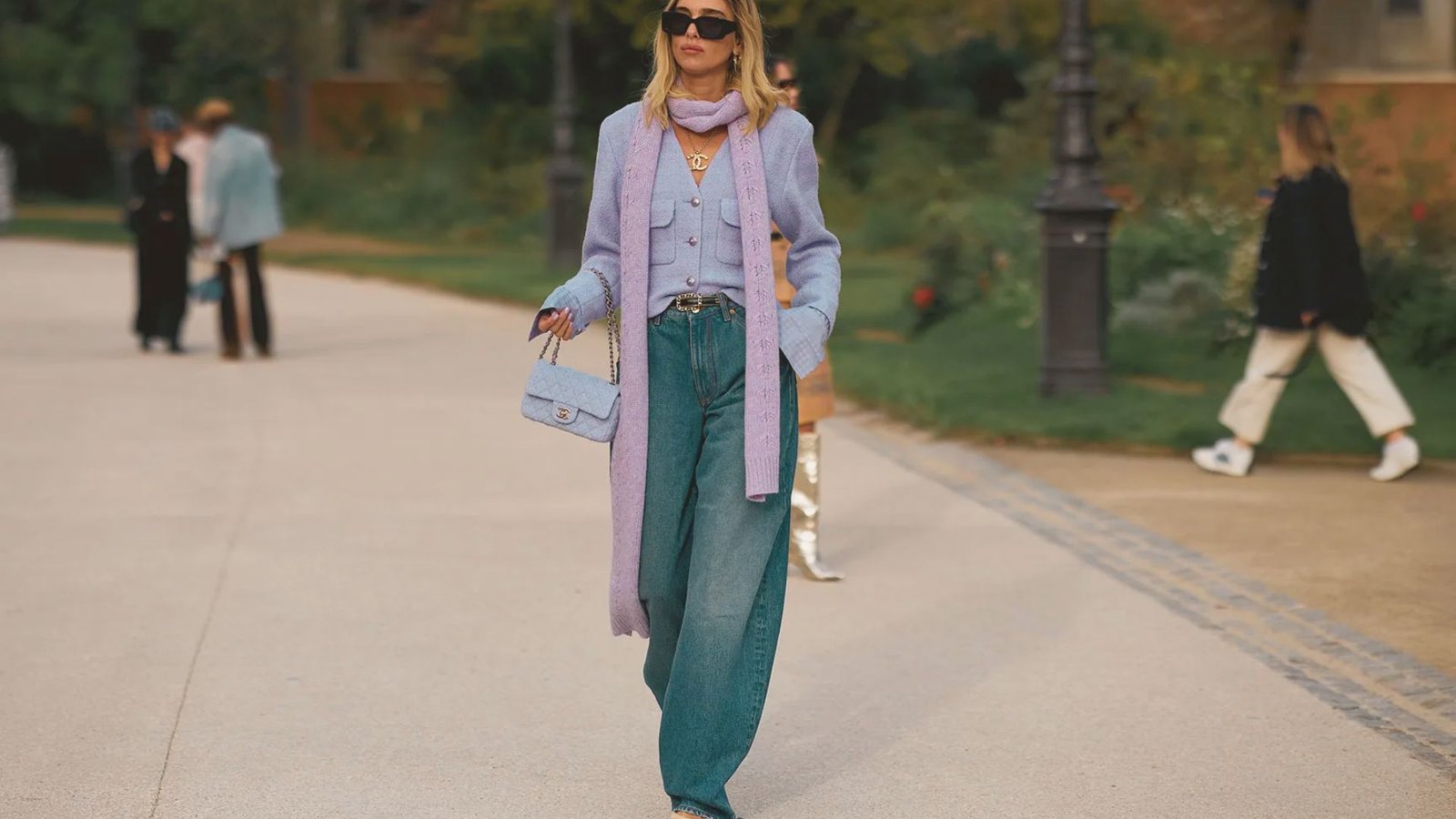 A woman walks confidently in a park wearing a stylish lavender coat, matching scarf, and blue jeans. She carries a light blue handbag, exuding elegance and modern fashion.