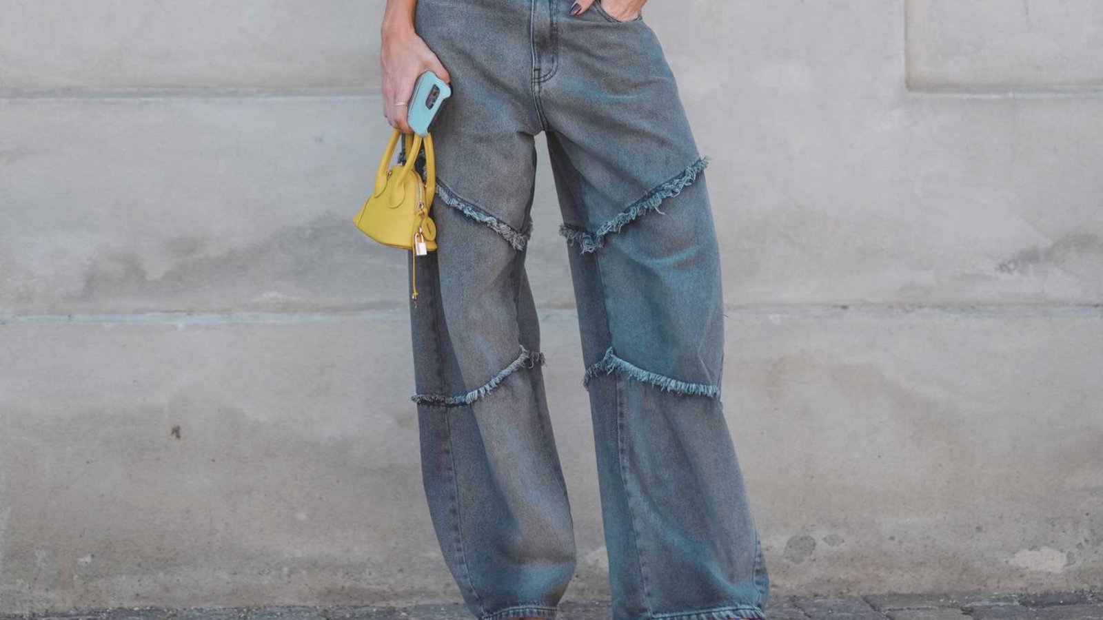 Wide-legged jeans with frayed seams are paired with a yellow mini handbag and a phone in hand against a neutral wall, creating a casual, trendy vibe.