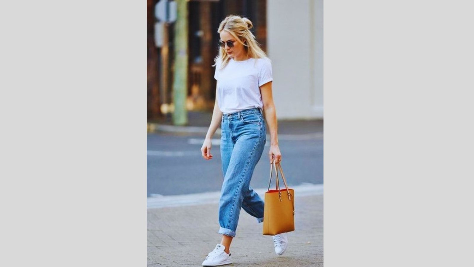 A woman in a white T-shirt and rolled-up jeans walks confidently on a street, holding a brown handbag. She wears sunglasses and white sneakers.