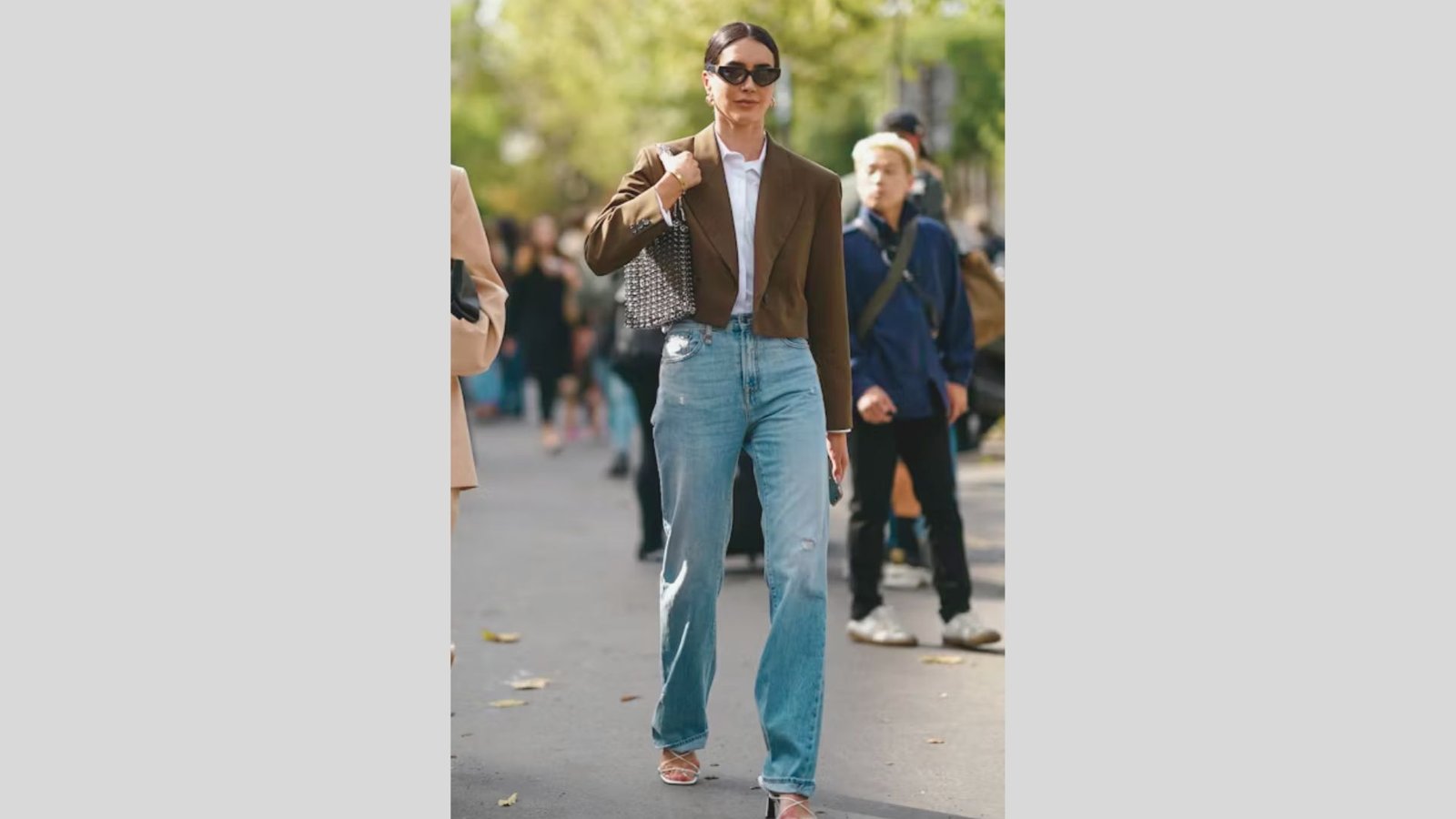 A woman in sunglasses walks confidently down a sunlit street, wearing a brown blazer, blue jeans, and pink heels, carrying a patterned bag.
