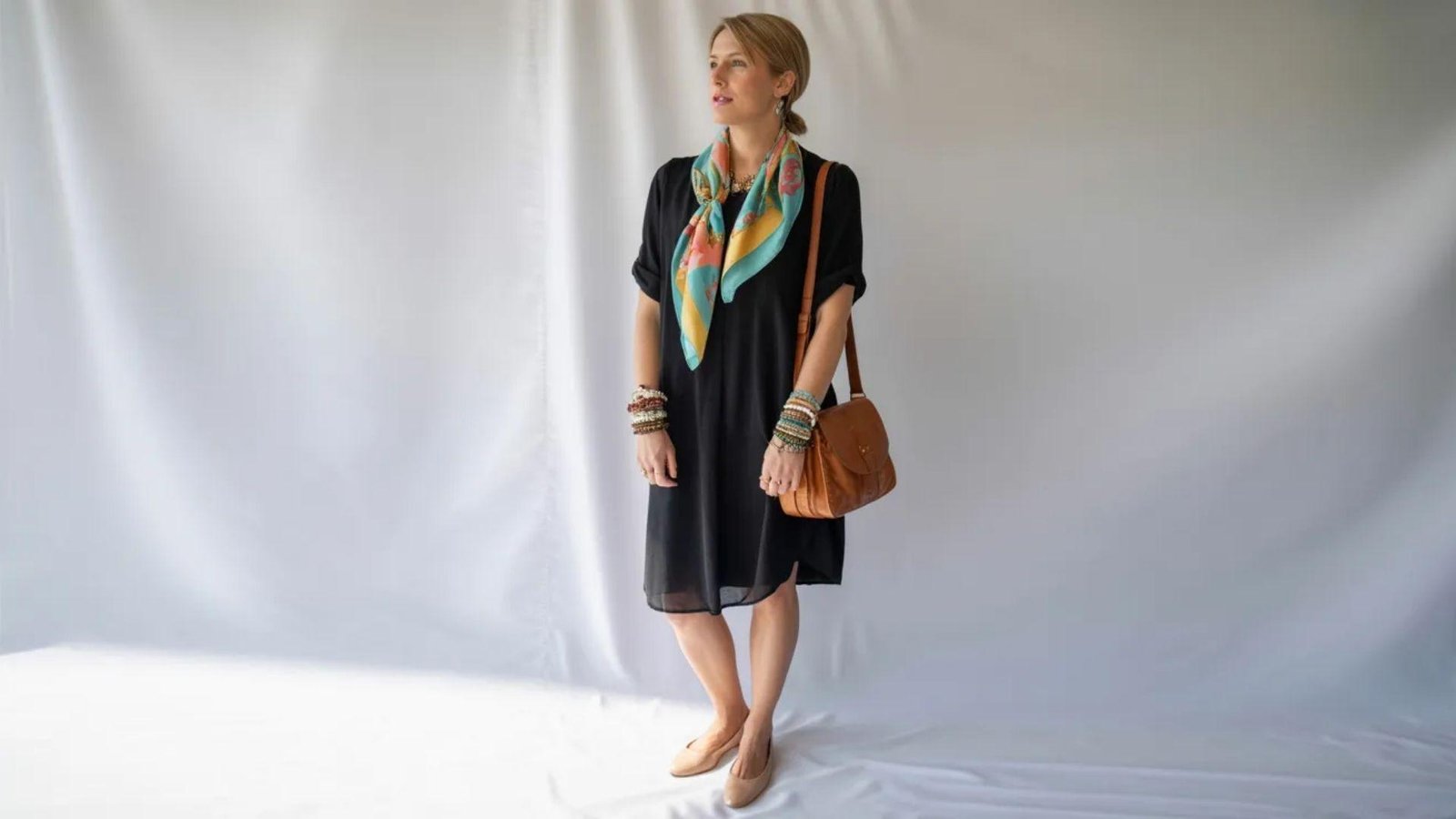 A woman wearing a black dress and scarf poses on a white background, highlighting her stylish and sophisticated appearance.