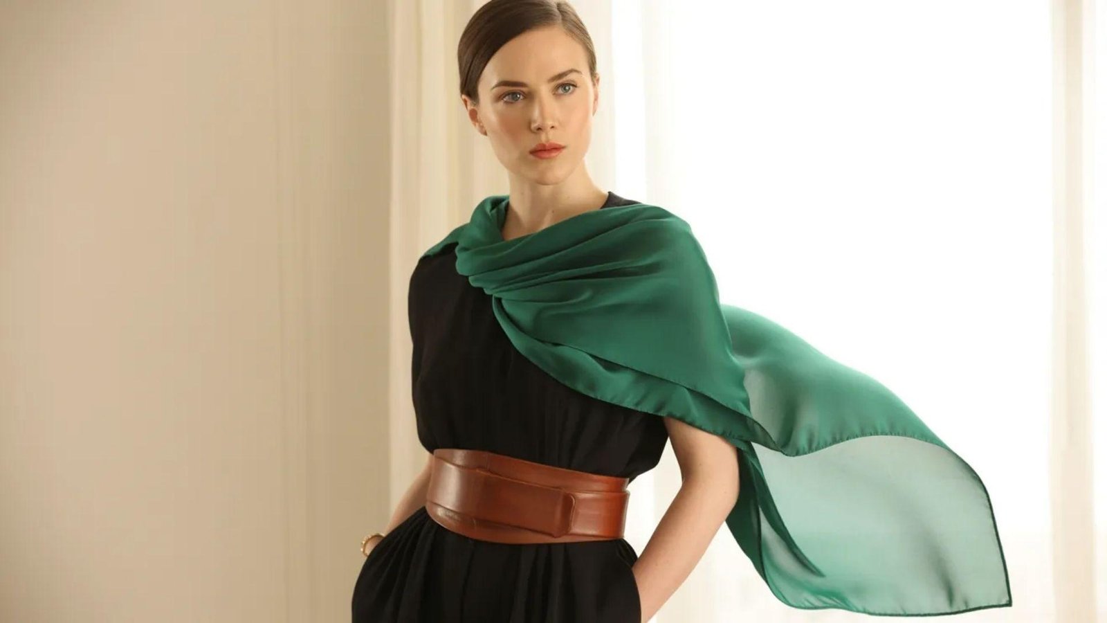 A woman wearing a black dress and a green scarf stands elegantly, showcasing her stylish outfit.