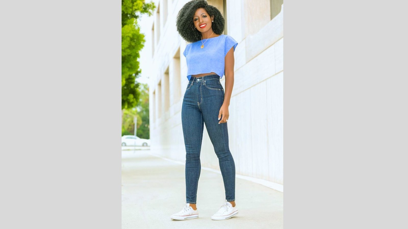 Woman in a blue top and high-waisted jeans stands confidently outdoors, smiling. She wears white sneakers; the setting is sunny with lush greenery.