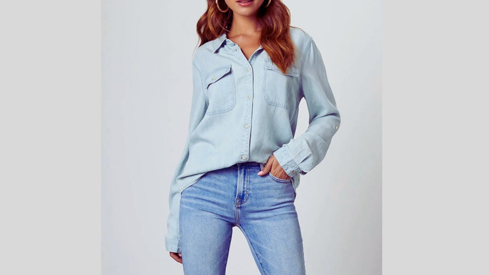 A woman in a light blue denim shirt and jeans stands against a white background. Her relaxed pose and casual attire convey a laid-back and stylish vibe.