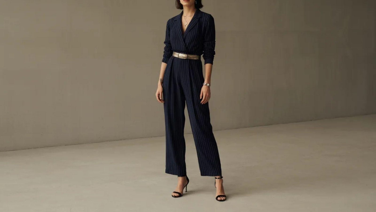 A woman wearing a navy jumpsuit and heels stands confidently, showcasing a stylish and modern outfit.
