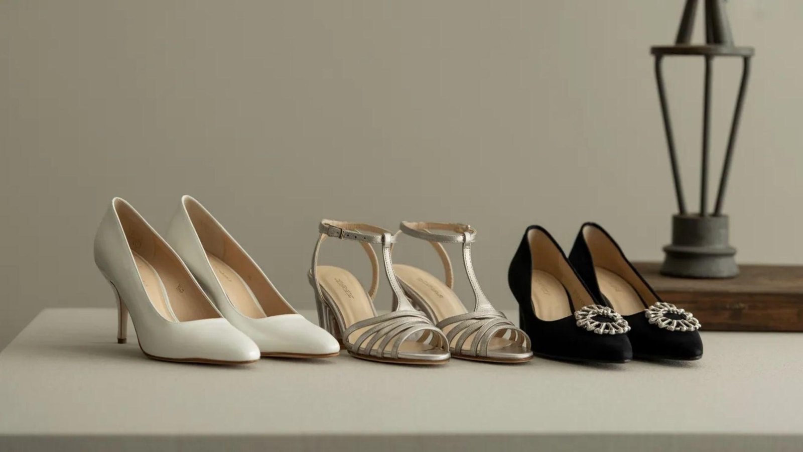 Three pairs of elegant women's shoes are displayed: white heels, silver strappy sandals, and black heels with jeweled accents, on a table with a neutral backdrop.