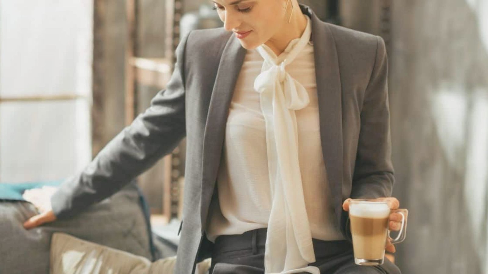 A woman in a gray suit with a white blouse sits on a couch holding a latte, exuding a calm, professional demeanor in a cozy, modern setting.