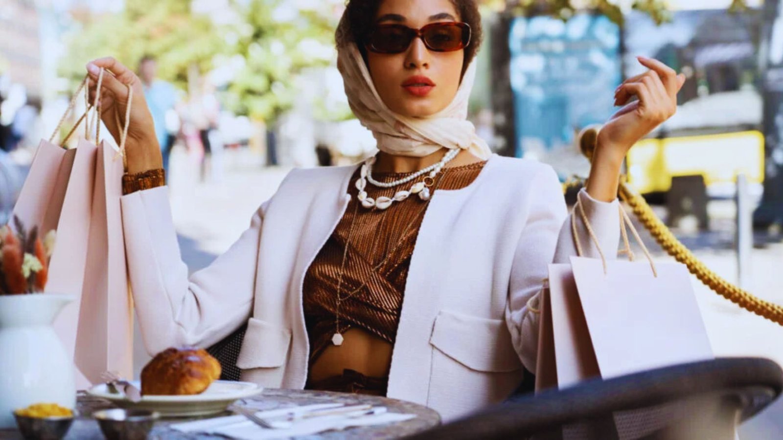 A woman in a headscarf sits at a table surrounded by shopping bags, smiling and enjoying her time.