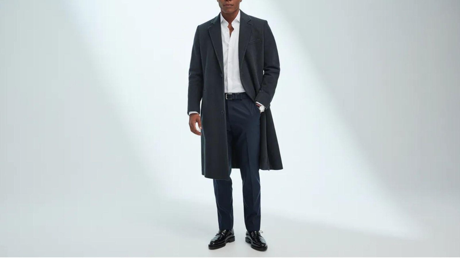A man wearing a long coat and pants stands confidently, showcasing a stylish and sophisticated look.