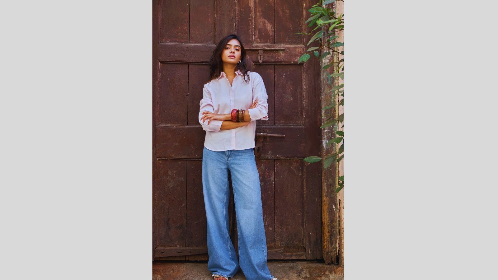 A person stands confidently in front of a rustic wooden door, wearing a light shirt and wide-legged jeans. The scene conveys a relaxed, earthy vibe.