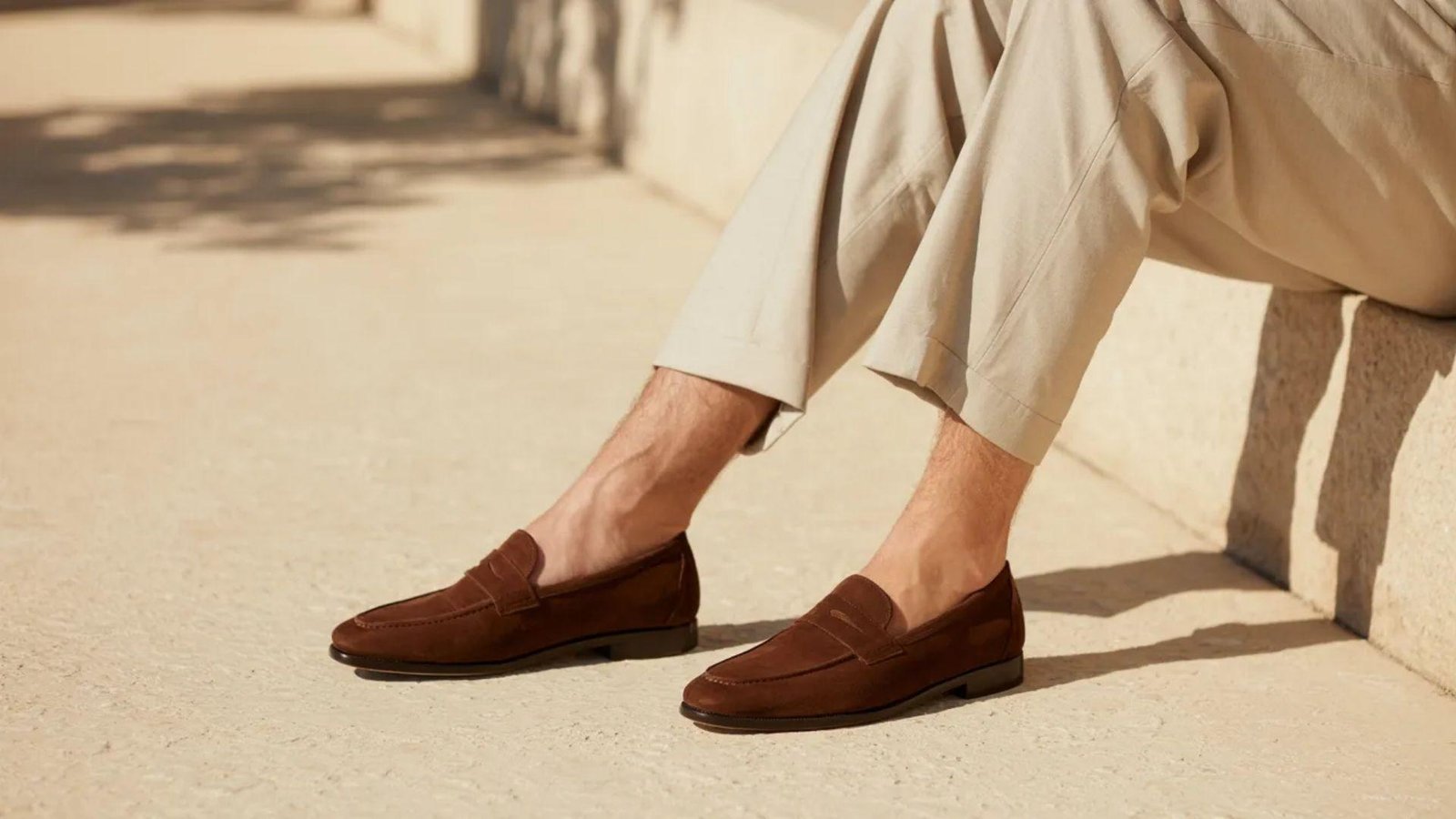 A man in brown shoes sits on a bench, looking relaxed in a casual outdoor setting.