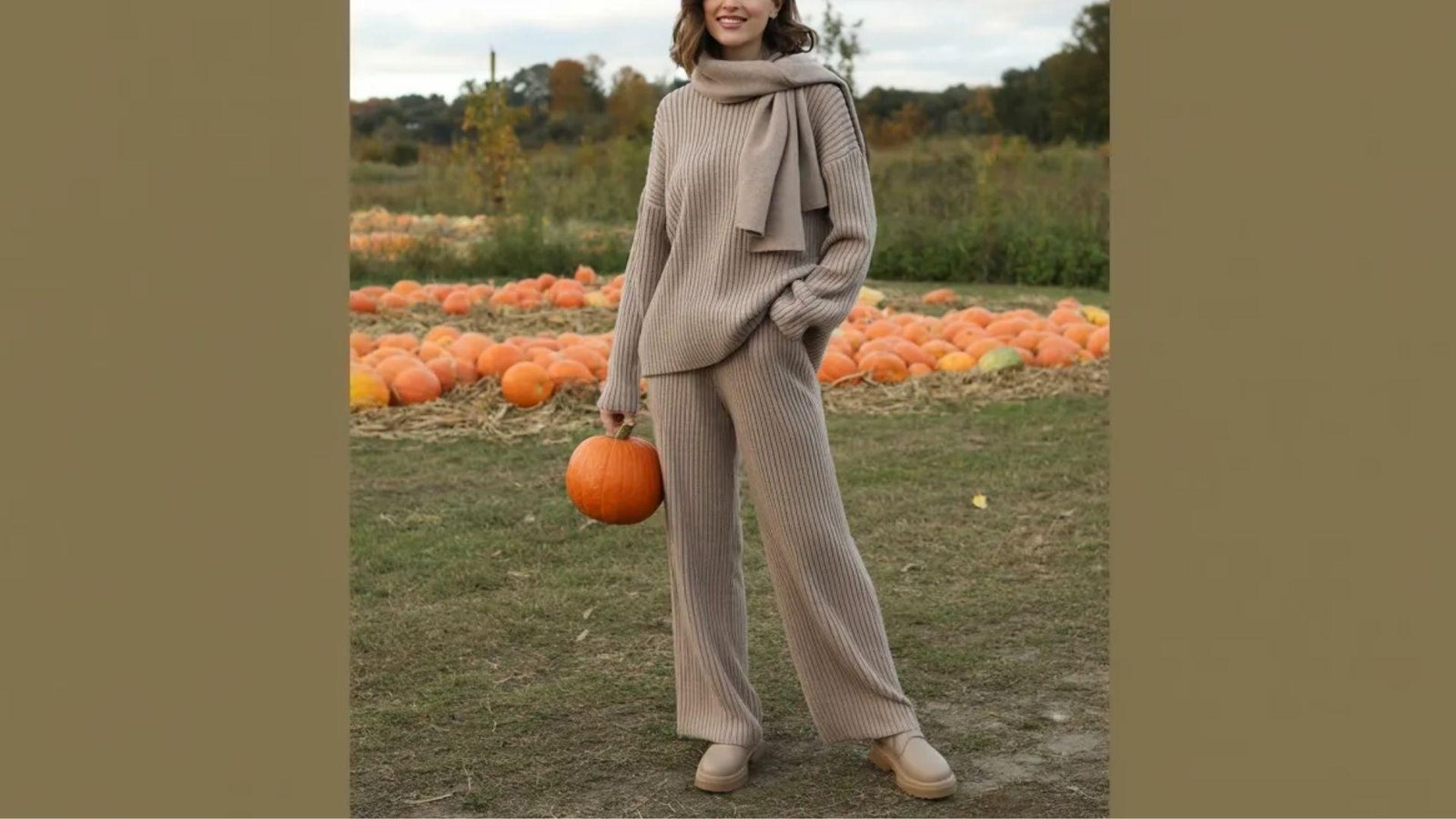 A woman wearing a sweater and pants holds a large pumpkin, smiling as she stands outdoors in a fall setting.