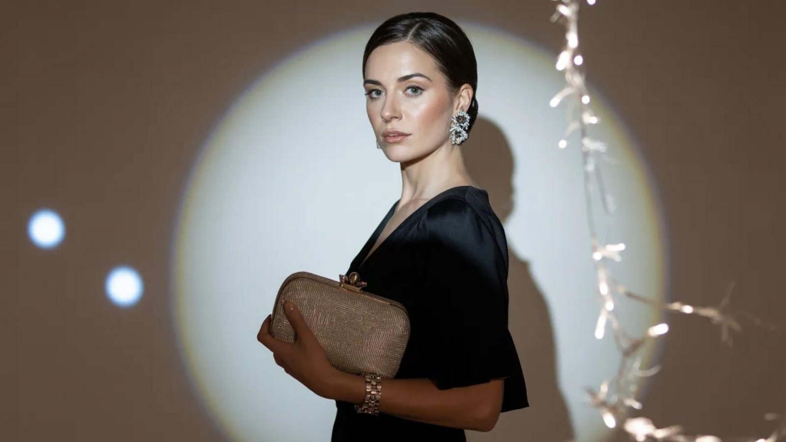 A woman in black attire holds a purse, standing in front of a bright light source.