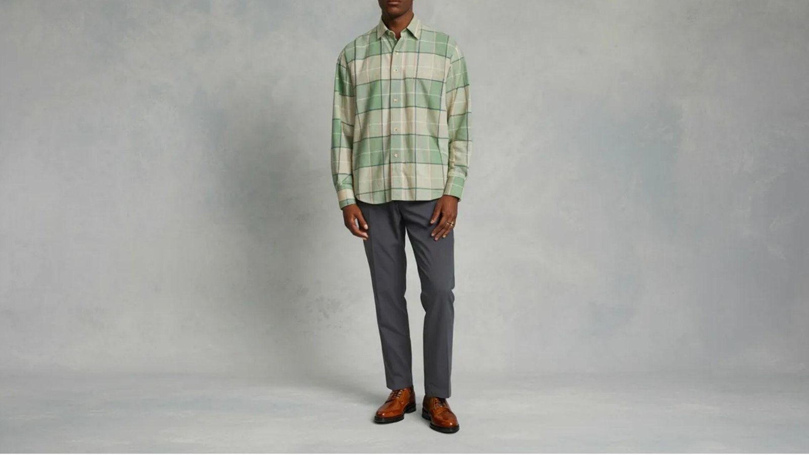 A man wearing a green and white checked shirt stands confidently, showcasing a casual yet stylish look.