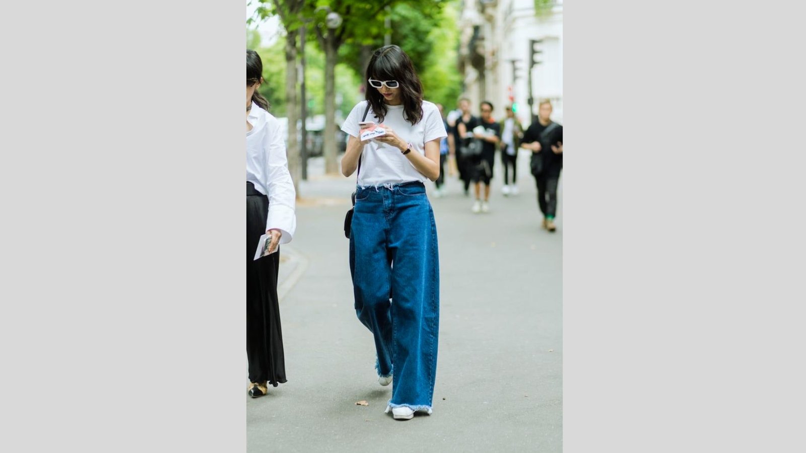 Woman walking down a tree-lined street, wearing white sunglasses, a white t-shirt, and loose blue jeans, looking at her phone. Casual, modern vibe.