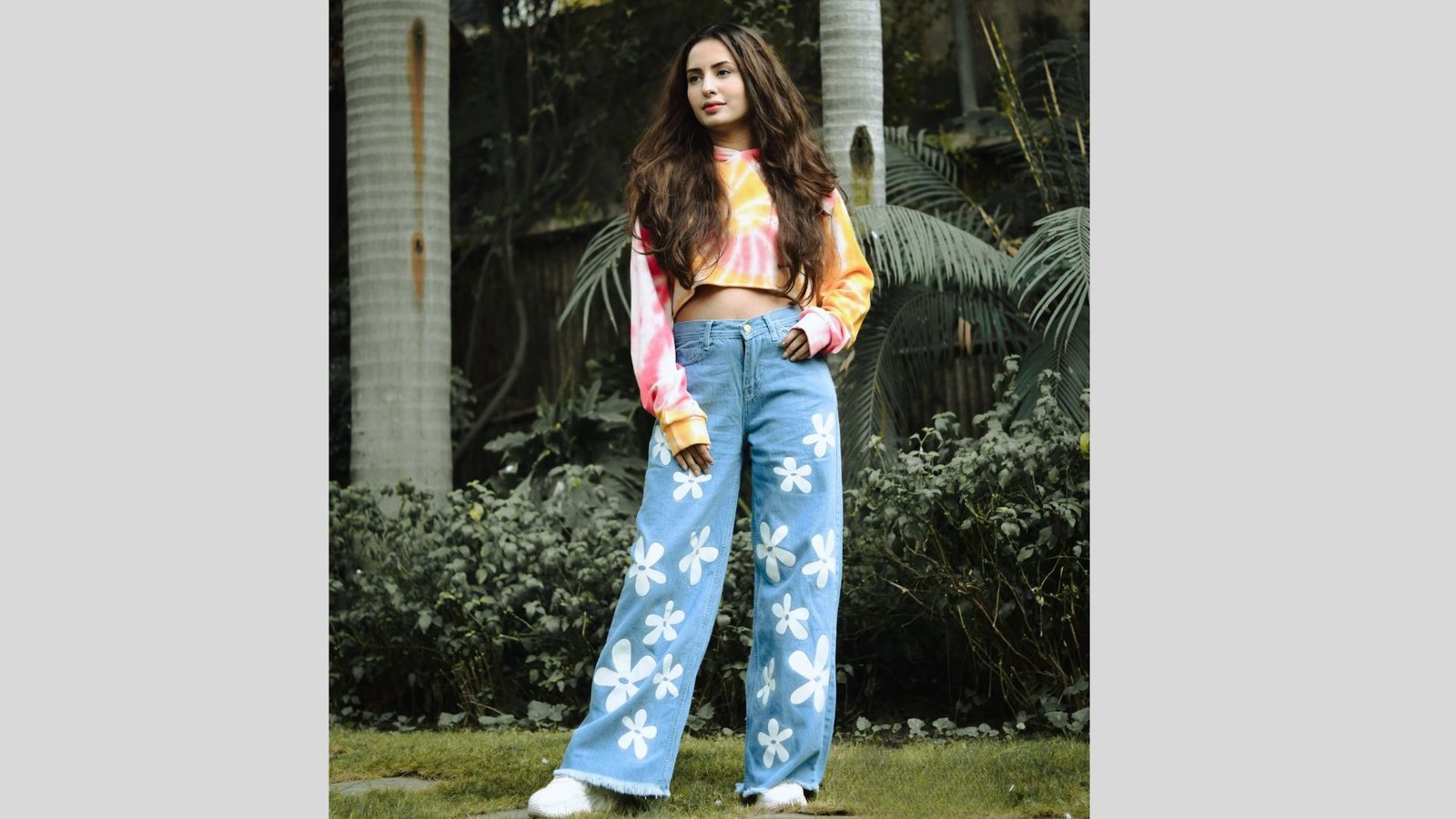 A woman stands on grass, wearing a colorful tie-dye crop top and blue jeans with white flower patterns. She looks confident amidst lush greenery.