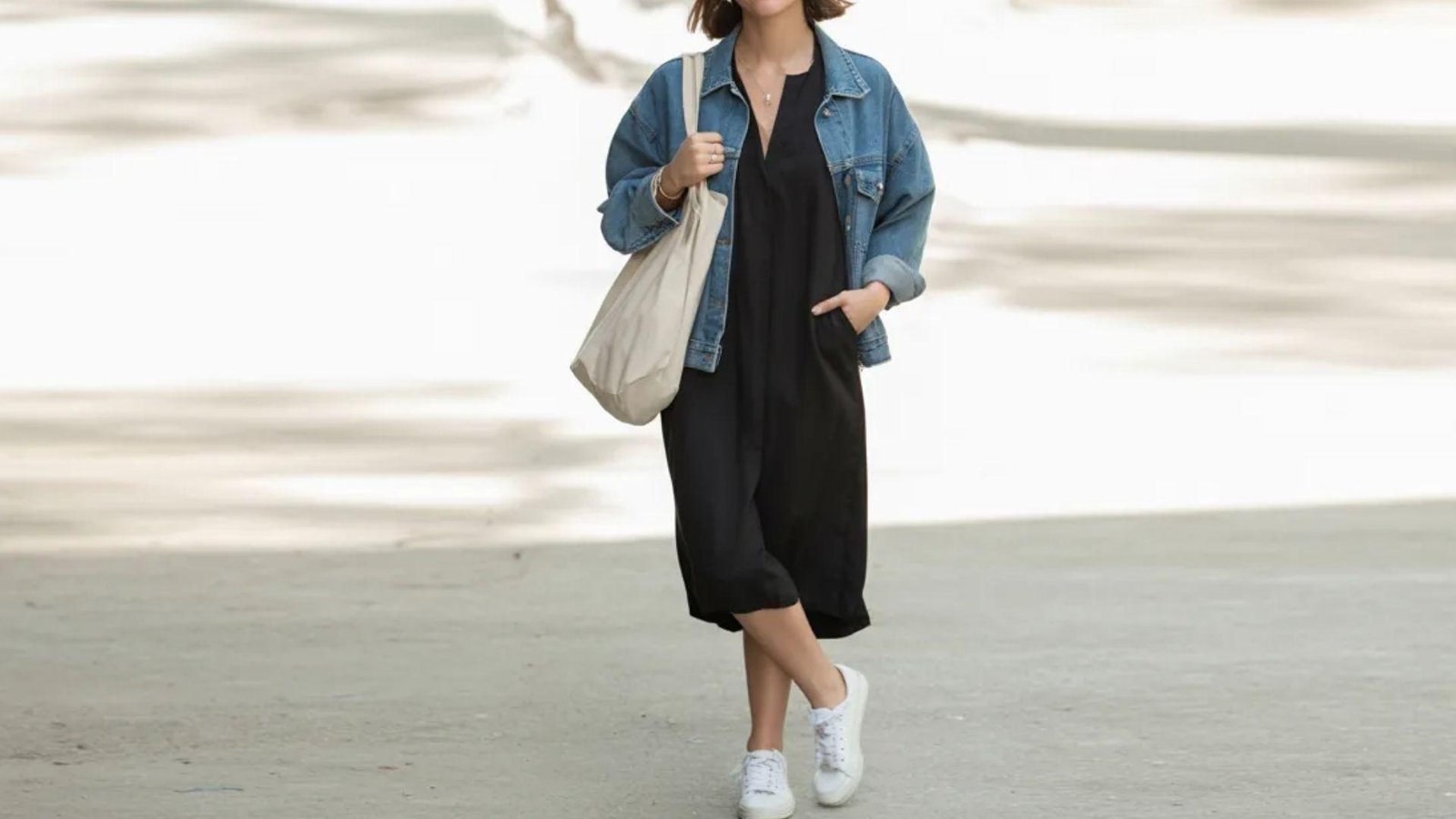A woman in a black dress and denim jacket walks confidently down the street.