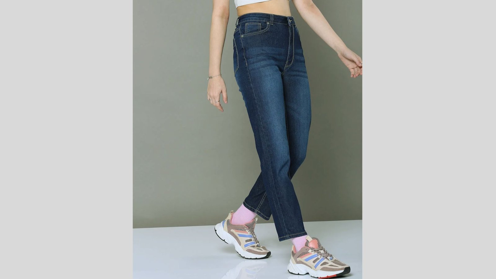 Woman in dark blue jeans and white sneakers with pink accents and pink socks, mid-stride. Her crop top is partially visible against a gray background.