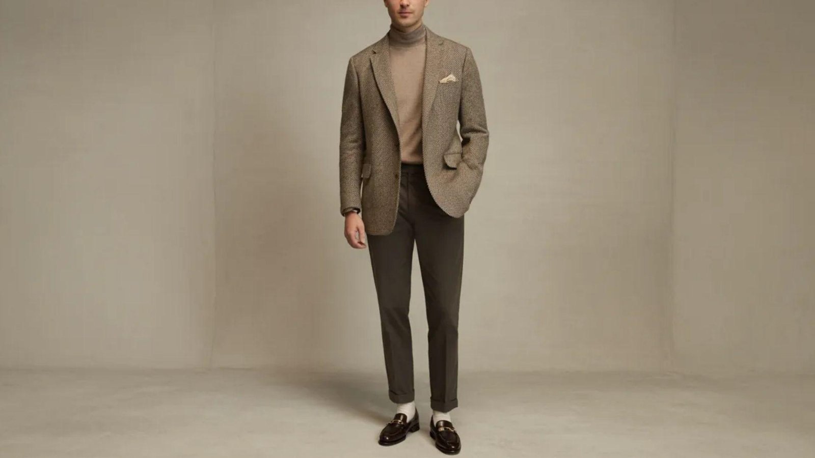 A man wearing a brown suit over a turtle neck sweater, standing confidently with a neutral expression.