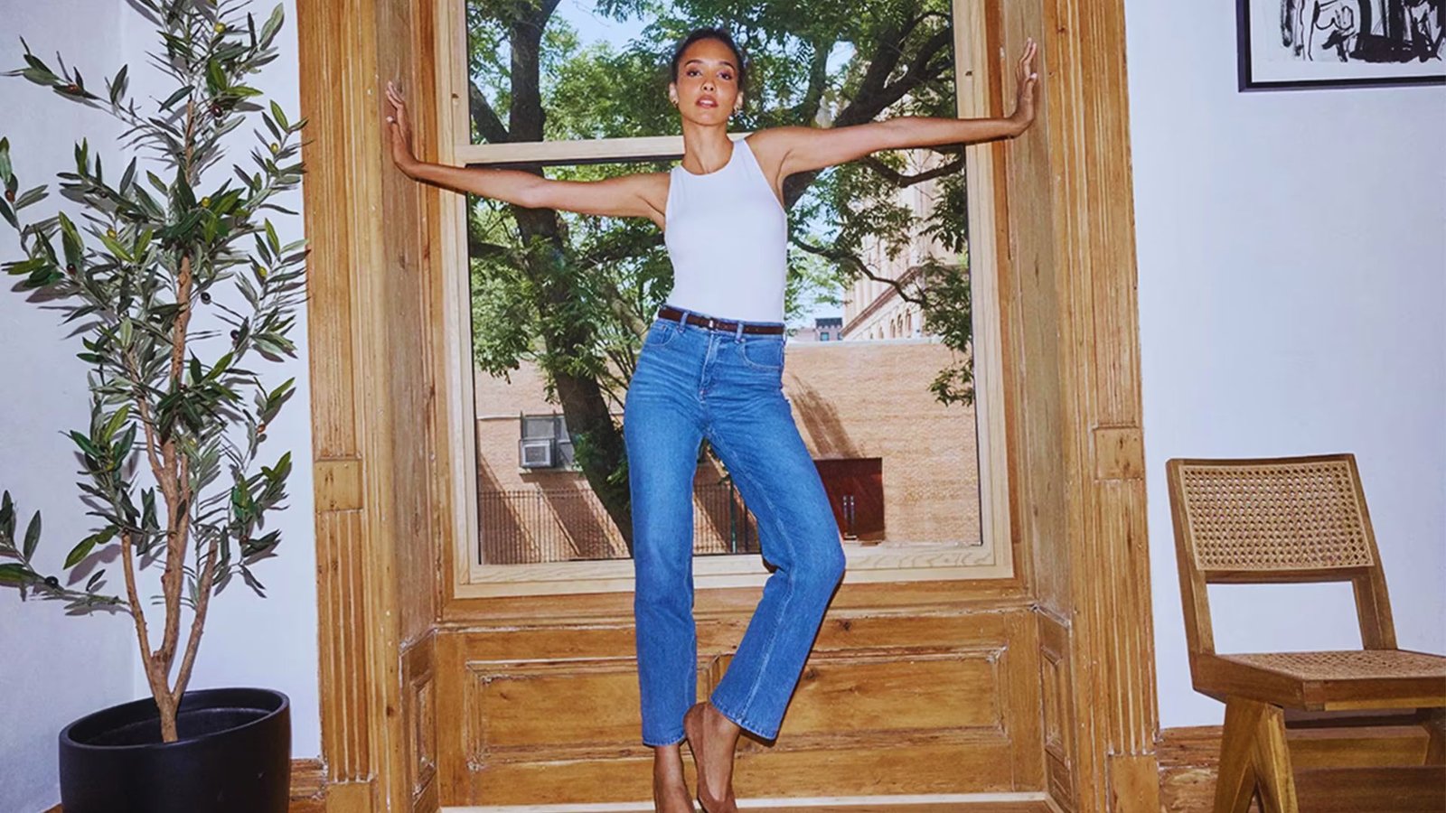 A person in a white tank top and blue jeans stands confidently in a sunlit wooden window frame. Greenery and a chair add to the room’s relaxed vibe.