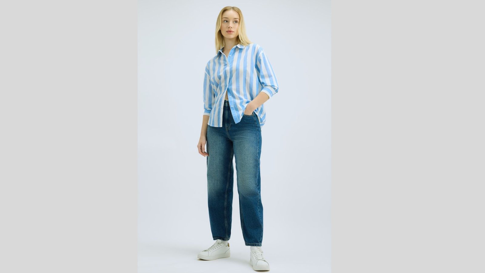 A person stands confidently wearing a blue and white striped shirt, blue jeans, and white sneakers; hands in pockets against a plain background.