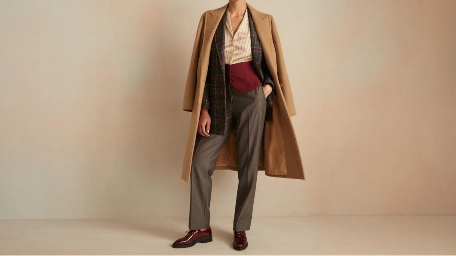 A person wearing a long beige coat over a checkered blazer and striped shirt, paired with gray pants and burgundy shoes, conveys a stylish, classic look.