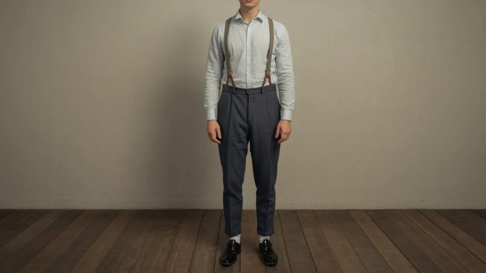 A man wearing suspenders stands on a wooden floor, showcasing a casual yet stylish appearance.

