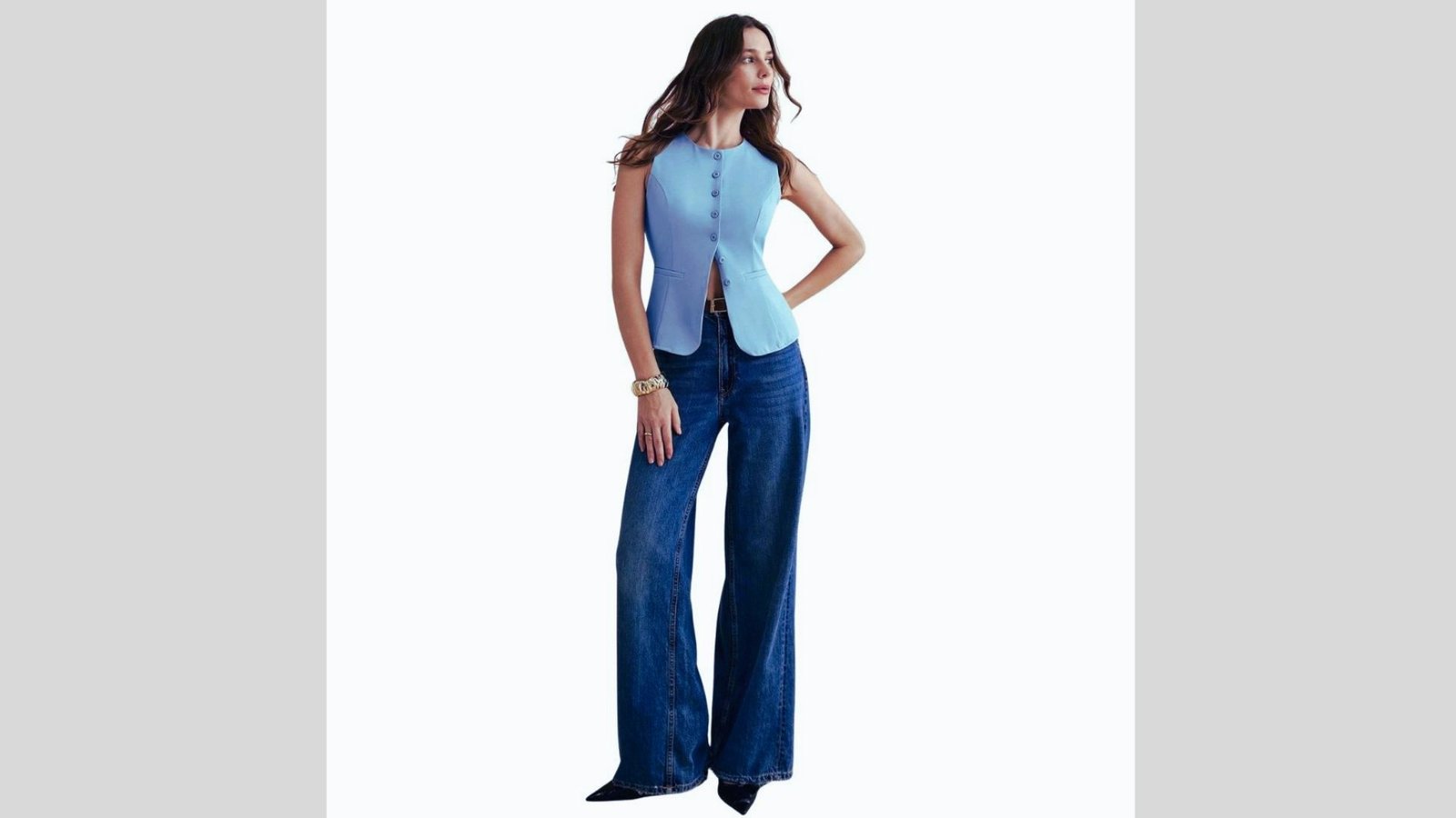 A woman stands confidently in a sleeveless blue top and wide-leg jeans against a plain background. Her relaxed pose and slight smile convey elegance.