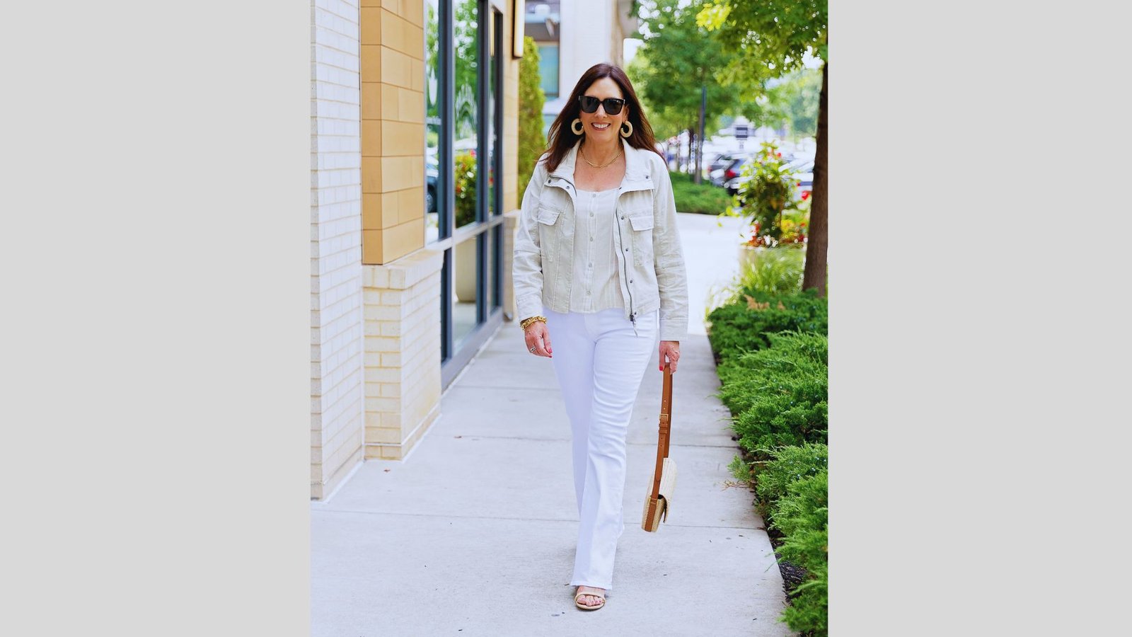 Woman walking confidently on a sidewalk, wearing sunglasses, a light jacket, white pants, and holding a handbag. Lush green trees in the background.