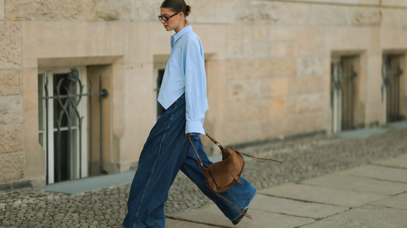 A woman walks confidently in wide-leg jeans and a light blue shirt, carrying a brown bag. She wears glasses and has her hair in a neat bun.
