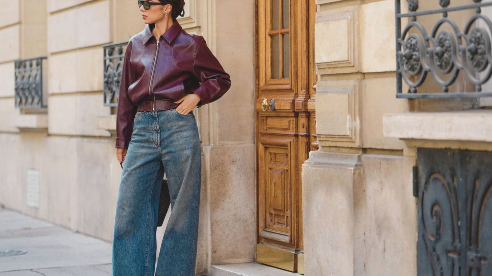 A woman in sunglasses stands confidently against a beige stone building, wearing a burgundy leather jacket and wide-leg jeans. The setting is stylish and urban.