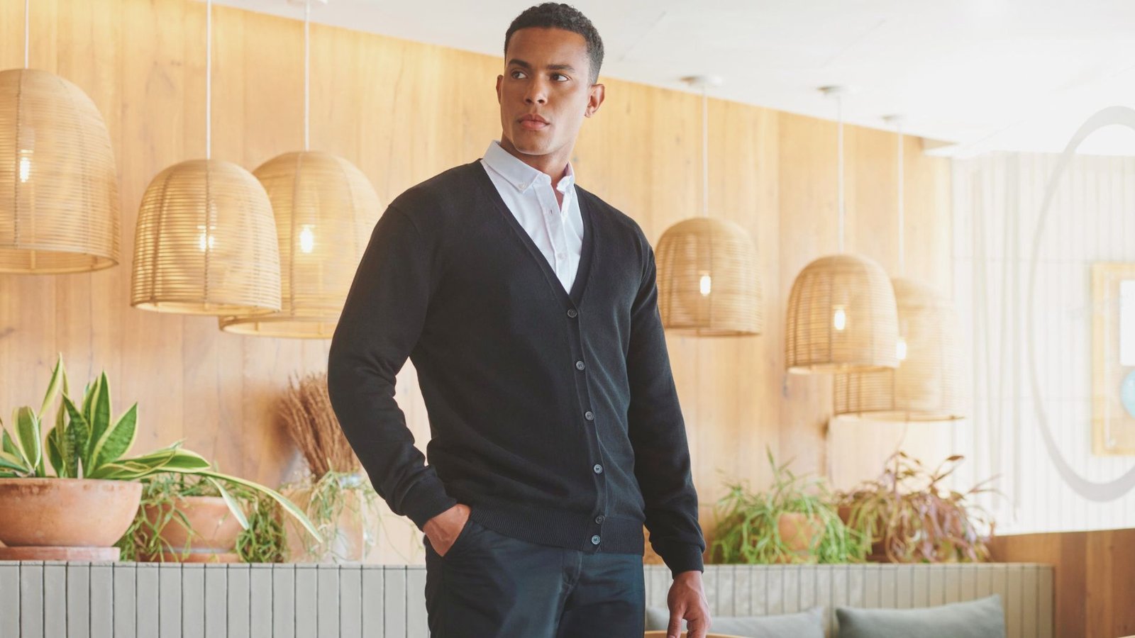 A man wearing a black cardigan and jeans stands in a room, looking towards the camera with a neutral expression.