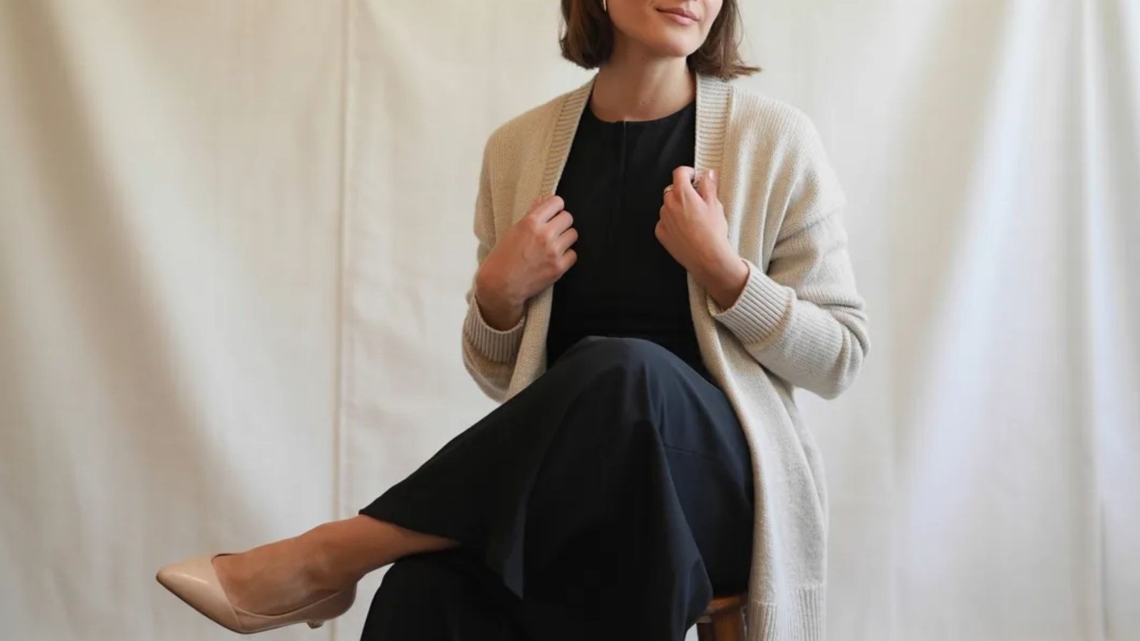 A woman wearing a black dress and a white cardigan stands confidently, showcasing a stylish and elegant outfit.