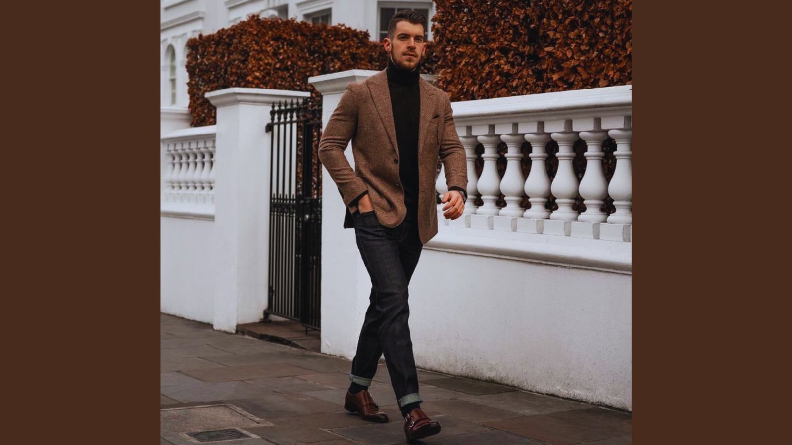 A man dressed in a brown blazer and black turtleneck strolls along the sidewalk.