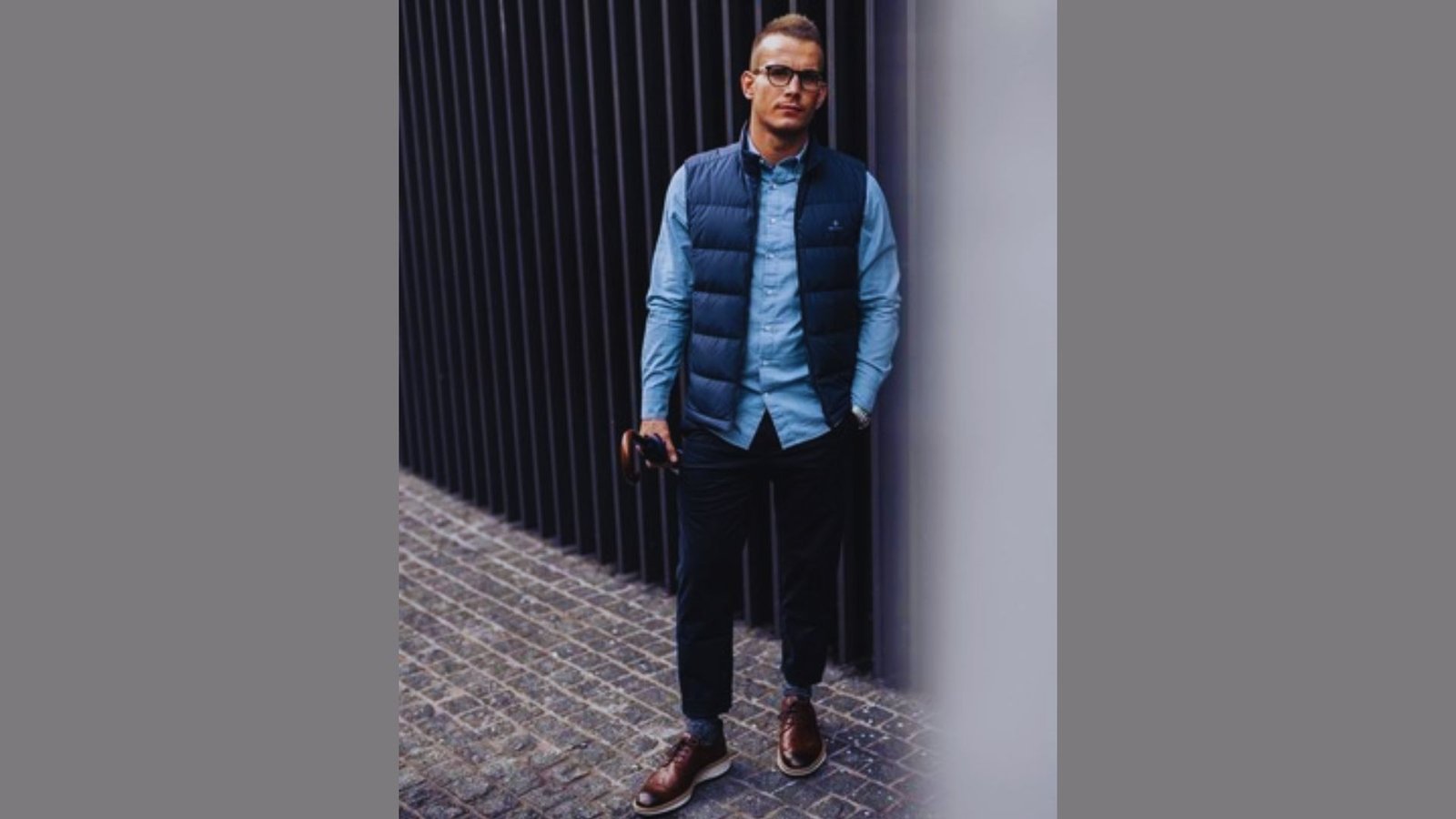 A man with glasses and a blue vest stands on a sidewalk, casually observing his surroundings.