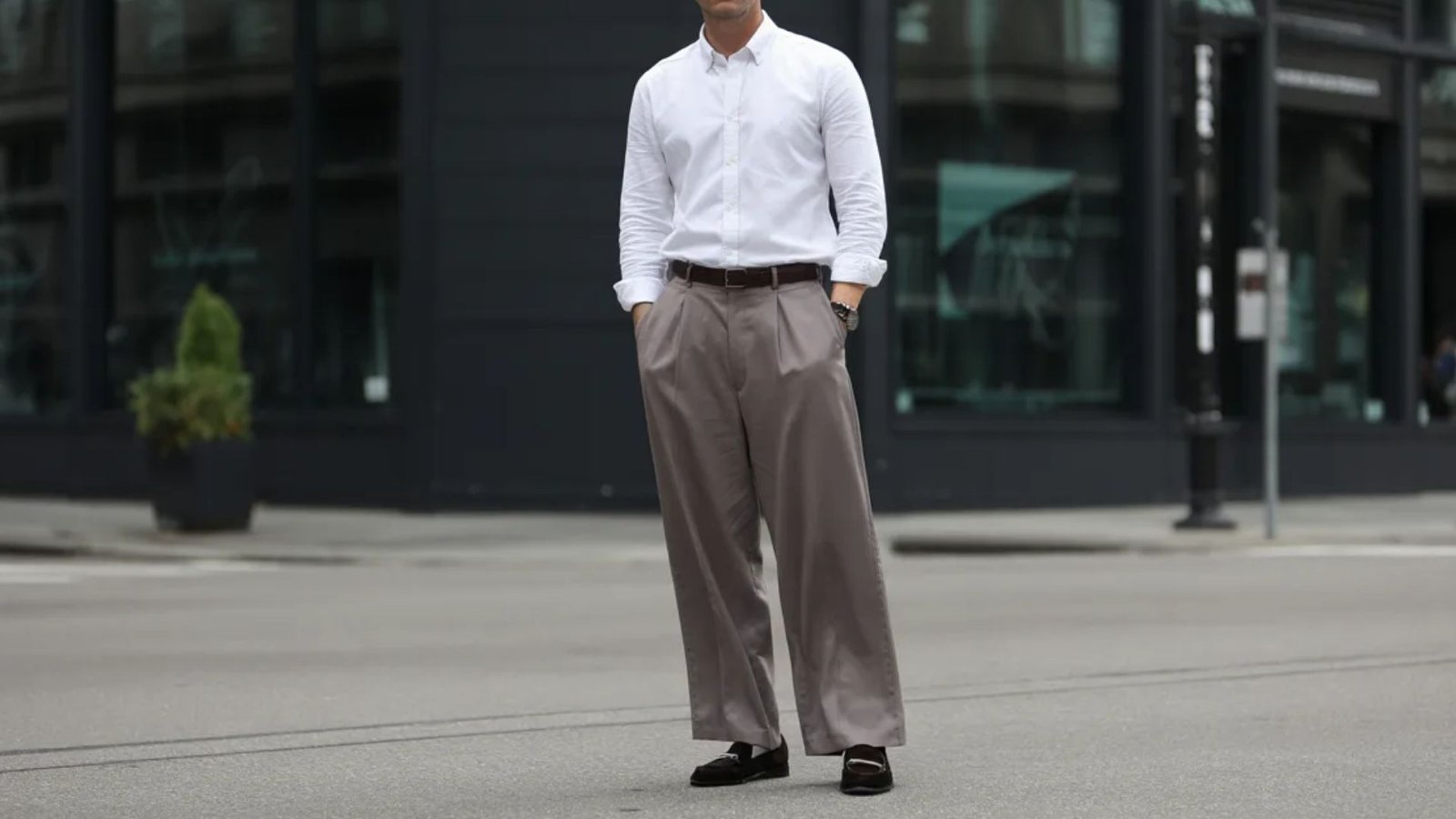 A man in a white shirt and slacks stands on the street, looking ahead with a neutral expression.
