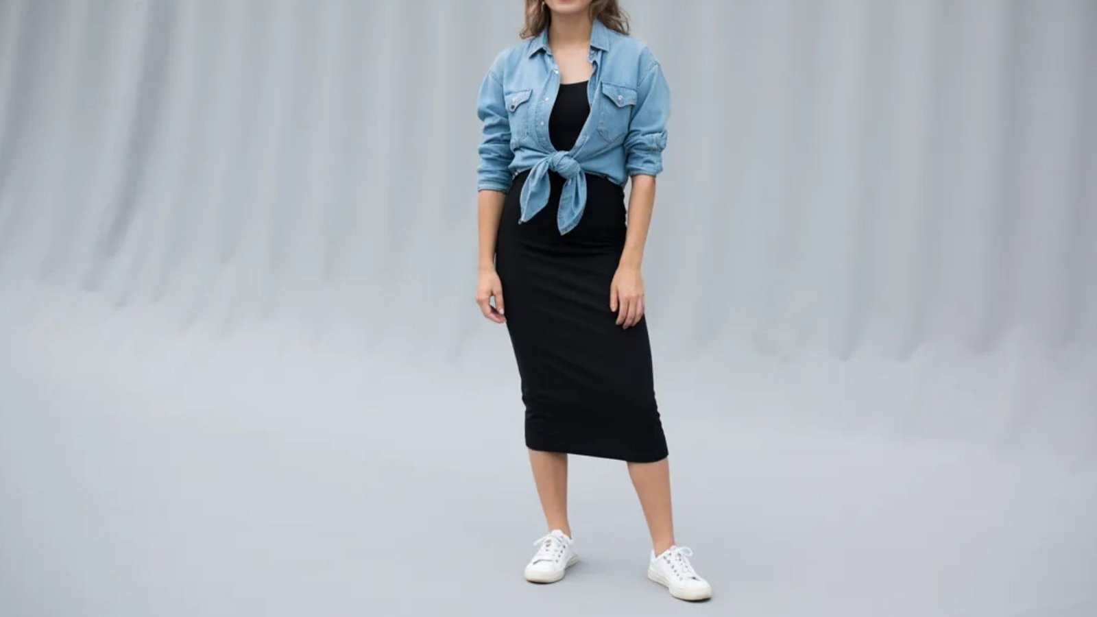 A woman wearing a black dress paired with a denim jacket stands confidently, showcasing a stylish casual look