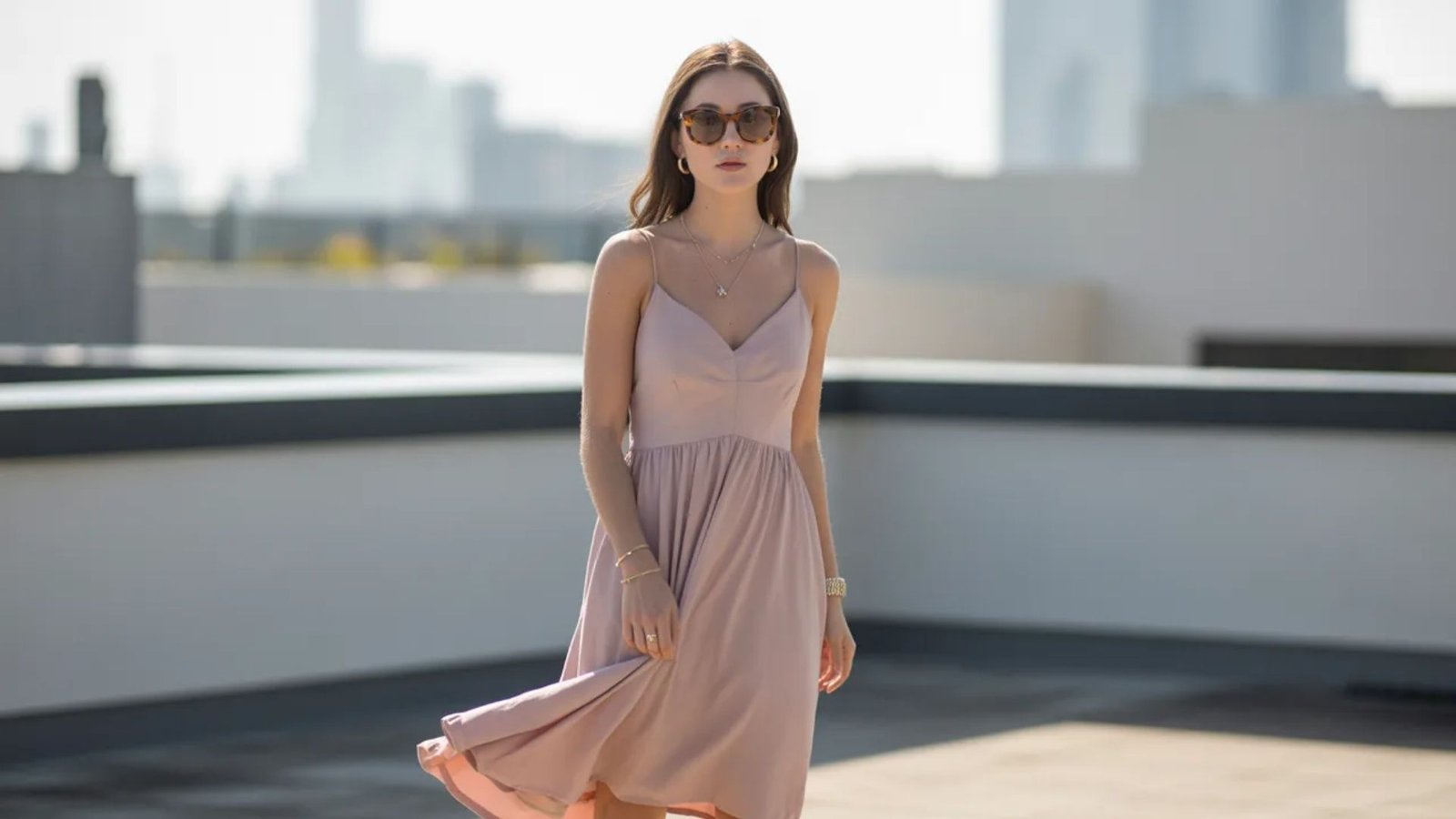  A woman in a pink dress and sunglasses strolls confidently on a rooftop, enjoying the urban skyline view.