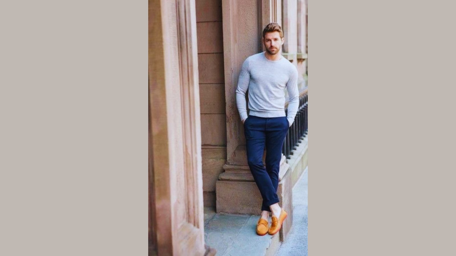 A man in a sweater and pants leans casually against a wall, looking relaxed and contemplative.