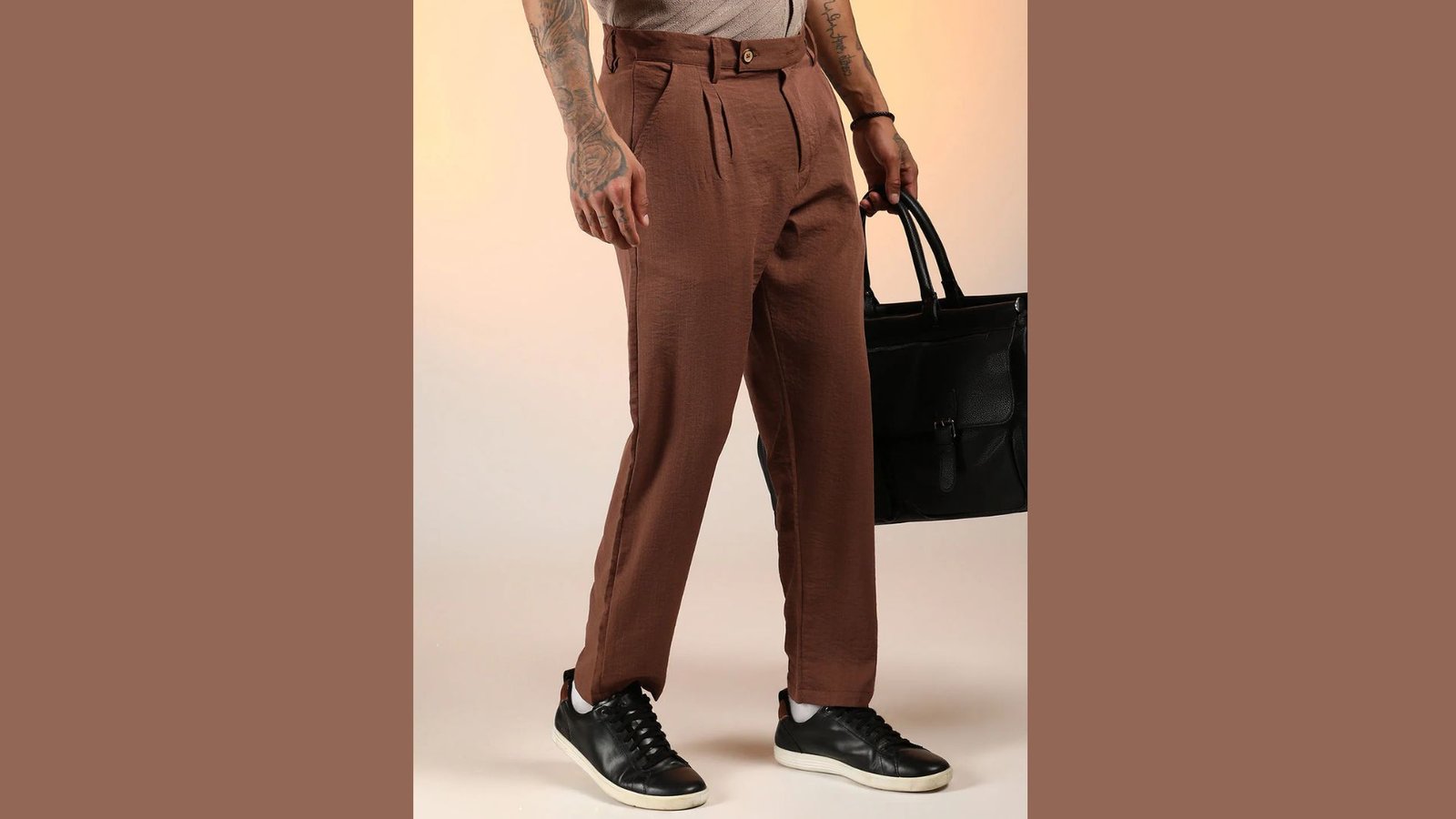 A man wearing brown pants and carrying a black bag stands confidently, showcasing a casual yet stylish look.