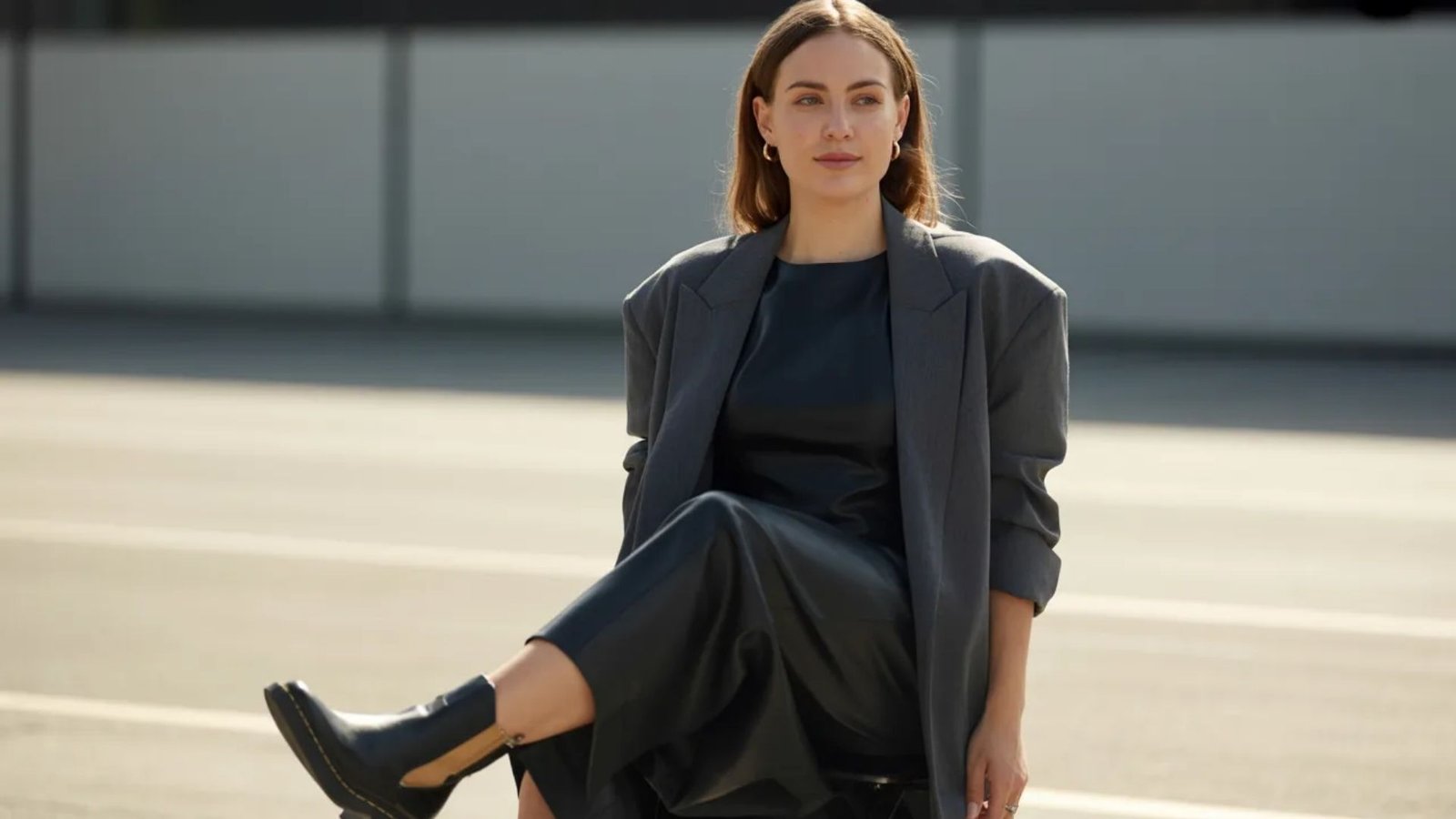 A woman wearing a black dress and leather boots sits on the ground, her posture relaxed and thoughtful.
