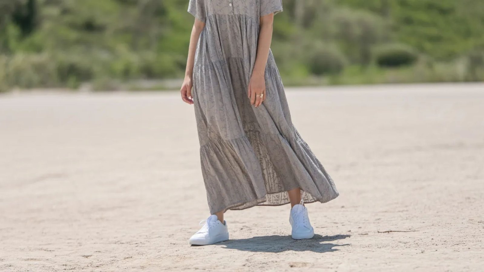 A woman in a long grey dress stands gracefully on the beach, with waves gently lapping at her feet.