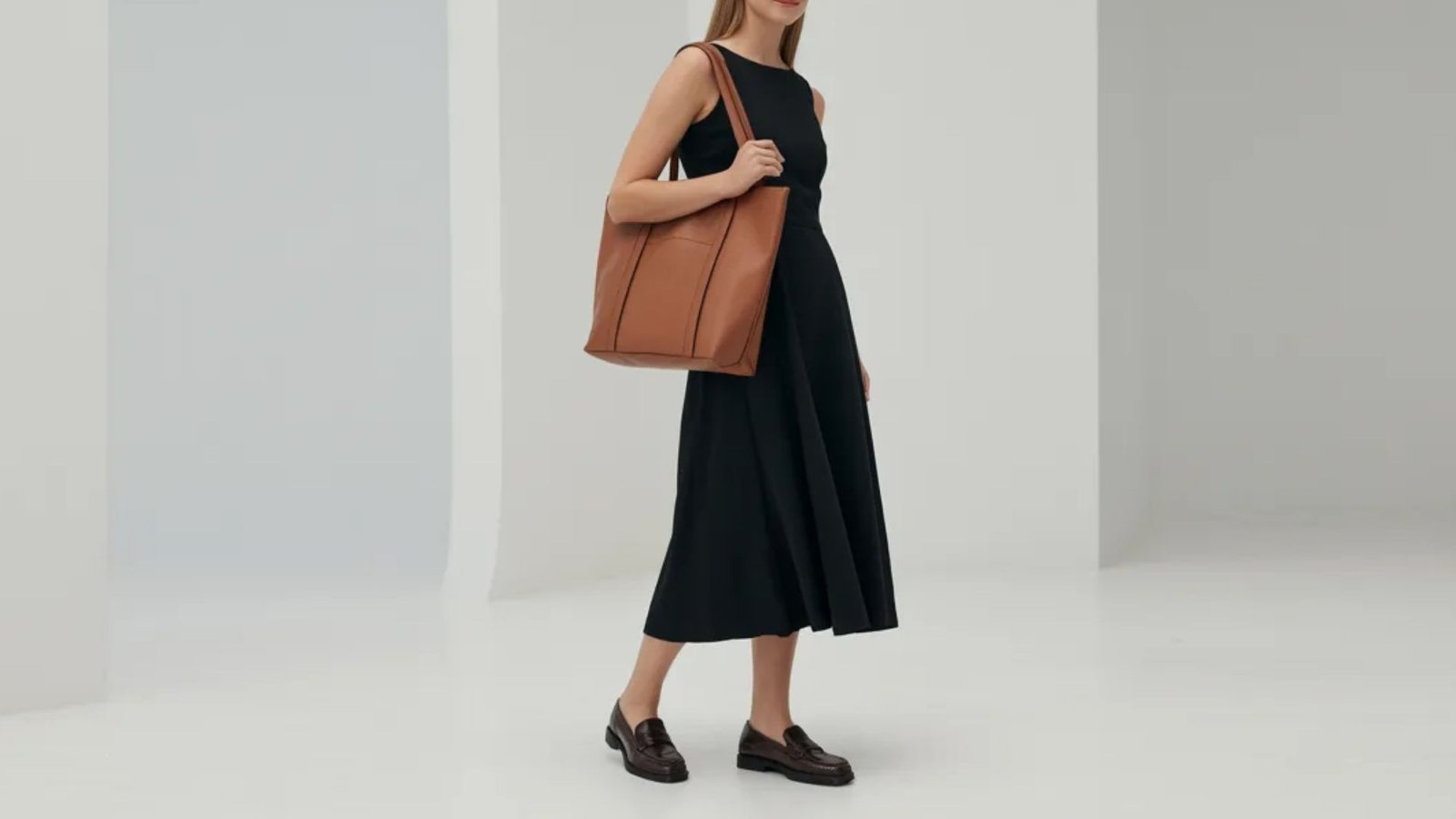 A model is dressed in a sleek black dress, accessorized with a brown handbag.