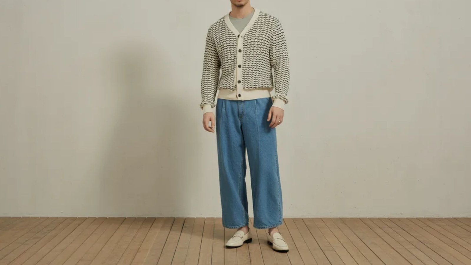 A man wearing a striped cardigan and blue jeans stands casually, showcasing a relaxed and stylish outfit.