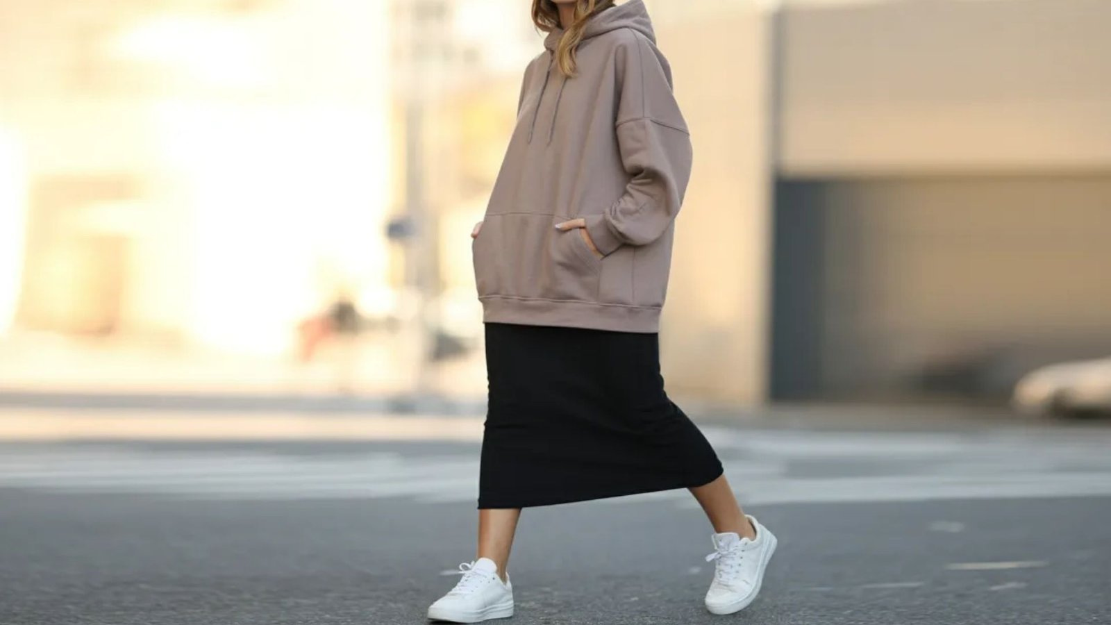 A woman in a long skirt and hoodie walks down the street, surrounded by urban scenery.