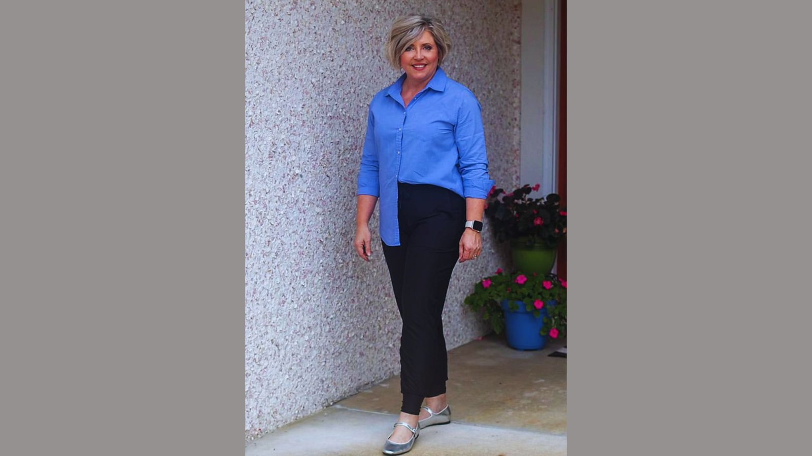 A woman wearing a blue shirt and black pants stands confidently, showcasing a casual yet stylish outfit.