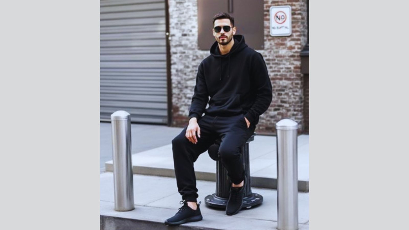 A man wearing a black sweatshirt sits on a curb, looking contemplative in an urban setting.