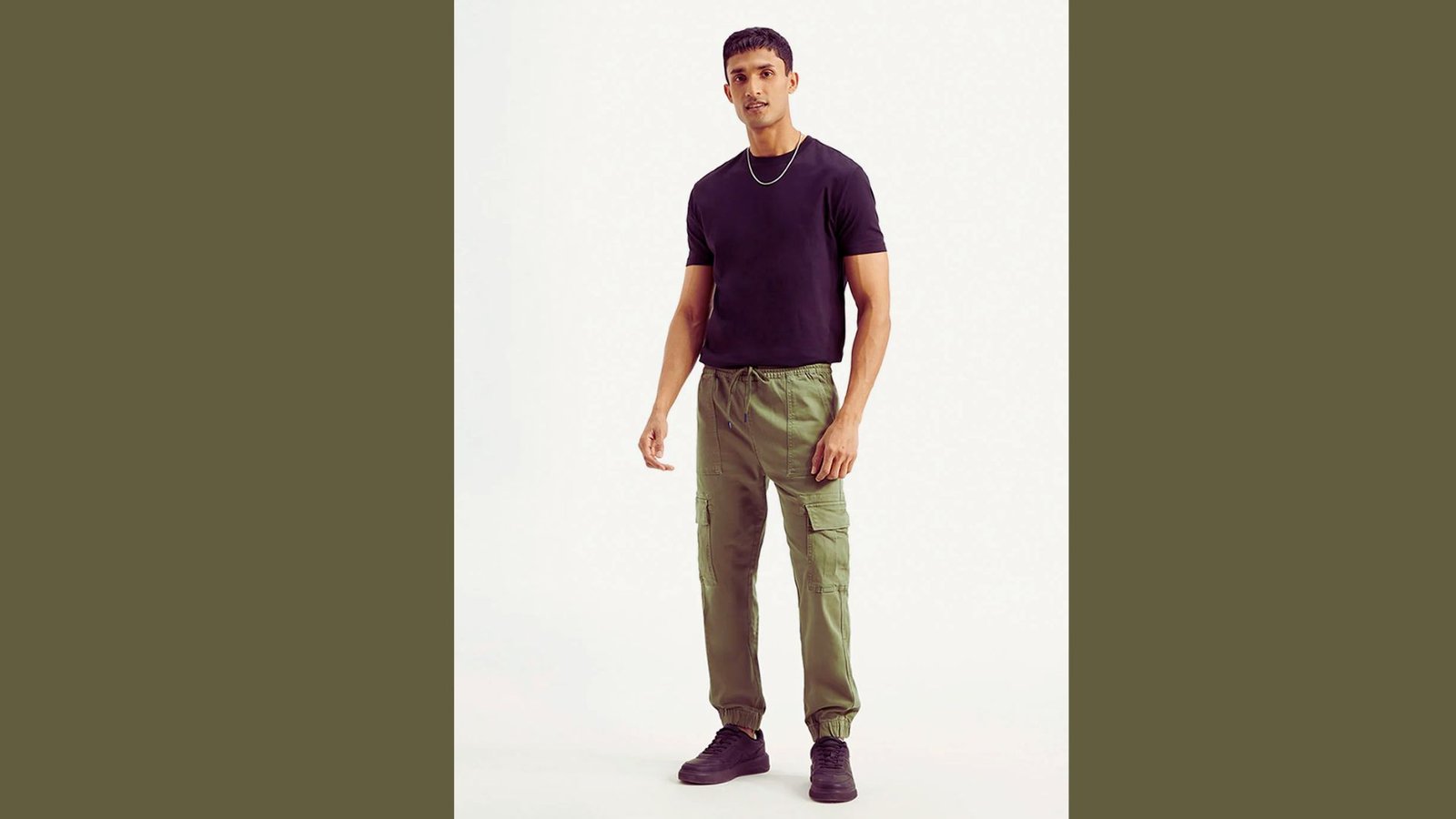 A man wearing a black shirt and green pants stands confidently, showcasing a casual yet stylish outfit.