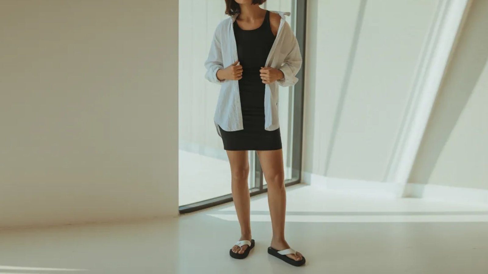 A woman in a black dress and white cardigan stands alone in a vacant room with plain walls and no furniture.